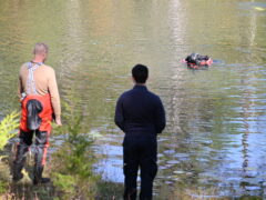White male wearing navy blue pants and sweatshirt standing beside another man wearing dive gear on the banks of a pond where a diver is off in the distance.