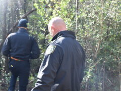 Two white men in long sleeve black jackets walk in a line in a wooded area.