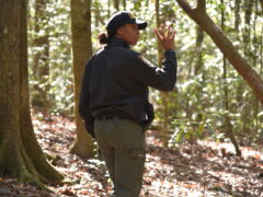 A black female deputy wearing a black ball cap and a black jacket holds up her hand with four fingers up as indicating the number four. She is standing in a wooded area.