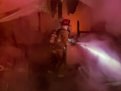 Firefighters with hose and flashlight inside a damaged home surrounded by smoke at night