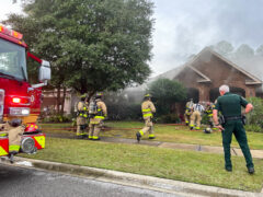 Firefighters, fire truck, and law enforcement officer outside of a brick home with smoke coming from the roof