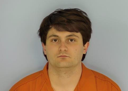Mug shot of a while male with brown hair and an orange jump suit frowning in front of a blue back drop.