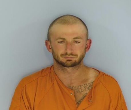 Mug shot of a while male with a balding head and brown beard.  The male is wearing an orange jumpsuit standing in front of a light blue wall. 