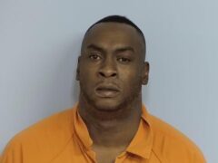 Mug shot of a black male with dark black hair and black eyes wearing an orange jump suit in front of a blue wall.