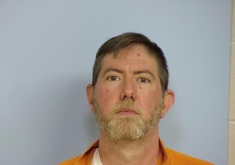 Mug shot of a white male with brown hair and a salt and pepper beard wearing an orange jump suit standing in front of a blue wall.