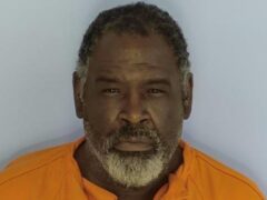 Mug shot of a black male squinting at the camera with an orange jumpsuit on standing in front of a light blue wall. He has salt and pepper beard with black hair.