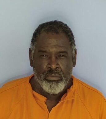 Mug shot of a black male squinting at the camera with an orange jumpsuit on standing in front of a light blue wall. He has salt and pepper beard with black hair.