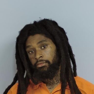 Mug shot of a black male with long think dreads wearing an orange jumpsuit standing in front of a light blue wall.