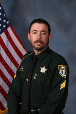 A white male dressed in a Class-A Deputy Sheriff Uniform standing in front of an American Flag