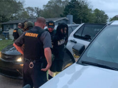 Two sheriff deputies, one in uniform, one in plain clothes with a bullet-proof vest on, arresting a black male with long thick black dreads putting him in the back of a patrol car.