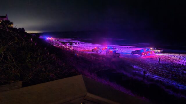 Deputy Sheriff vehicles lined up across the beach in front of a large crowd of teenagers at night.