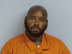Mug shot of a black male with a bald head and black beard wearing an orange jumpsuit in front of a light blue wall.