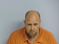 Mug shot of a bald white man with a gray and brown goatee wearing an orange jumpsuit standing in front of a light blue wall