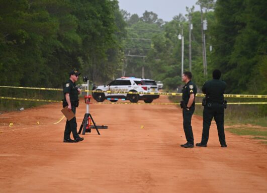Three deputies in uniform standing on a red dirt road with crime scene tape across it. 