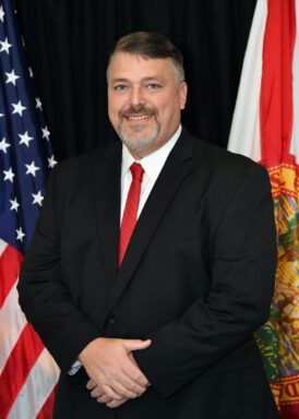 A photo of a white male with brown hair and a brown and gray beard wearing a suit with a red tie standing in front of the Florida State Flag and the American flag. 