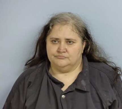 White woman with black hair and white roots wearing a navy blue jumpsuit standing in front of a light blue wall