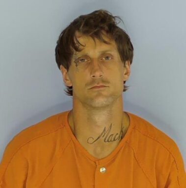 Mug shot of a white male with brown hair and a facial tattoo over the right eye and a tattoo on his neck reading "MACK" 