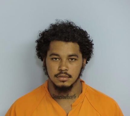 Mug shot of a black male with black hair and a black beard wearing an orange jumpsuit in front of a light blue wall.