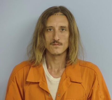 Mug shot of a white male with shoulder length dirty blonde hair with a mustache wearing an orange jumpsuit standing in front of a light blue wall.