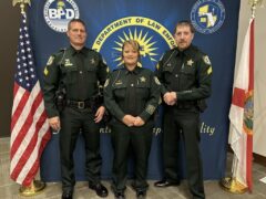 Three sheriff deputies, two men and one woman stand in front of the American flag and the Florida flag.