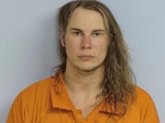 Mug shot of Shane Sanders, a white male with long brown hair passed his shoulders with an orange jumpsuit standing in front of a light blue wall.