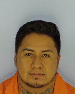 Mug shot of a Hispanic male with dark hair and short chin hair wearing an orange jump suit standing in front of a blue wall. 