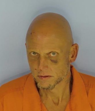 Mug shot of a bald white male with a bruise under his left eye wearing an orange jumpsuit standing in front of a light blue wall