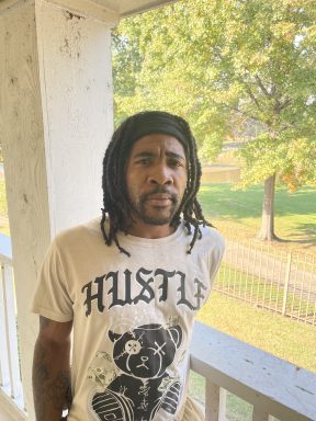 Photo a a black male with dreads and a black headband in handcuffs wearing a beige shirt that says "Hustle". 