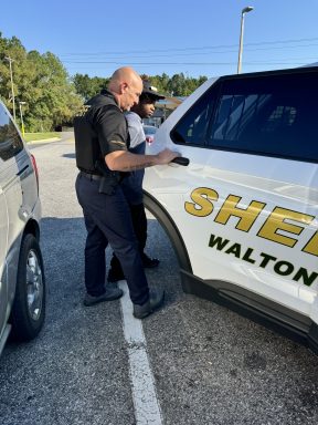 A deputy putting handcuffs on a black male teenager next to a patrol car.