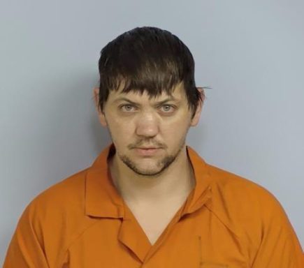 Mug shot of a white male with dark brown hair wearing an orange jumpsuit standing in front of a light blue wall