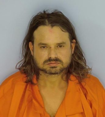 Mug shot of a white male with long brown hair and brown goatee wearing an orange jumpsuit standing in front of a light blue wall