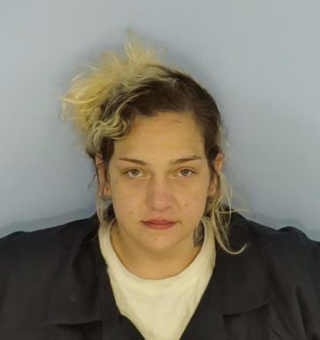 Mug shot of a white female with bleached blonde hair and dark brown roots wearing a navy blue jumpsuit and a white undershirt standing in front a light blue wall