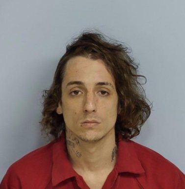 Mug shot of a white male with shoulder length brown hair and neck tattoos wearing an orange jumpsuit standing in front of a light blue wall.