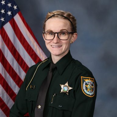 A photo of a female deputy with blonde hair and glasses sitting in front of an American flag