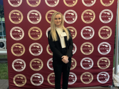 A blonde female with a black suit and white high heels standing in front of an FSU step and repeat.