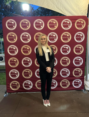 A blonde female with a black suit and white high heels standing in front of an FSU step and repeat.