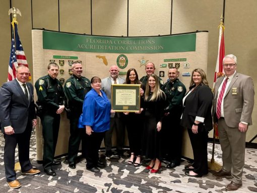 Group of deputies and non-sworn employees of the Walton County Sheriff's Office standing in front of a Florida Corrections Accreditation Commission banner holding the Excelsior Accreditation plaque