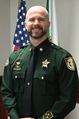 White male with dark brown beard wearing a dark green Class A Sheriff's Office uniform