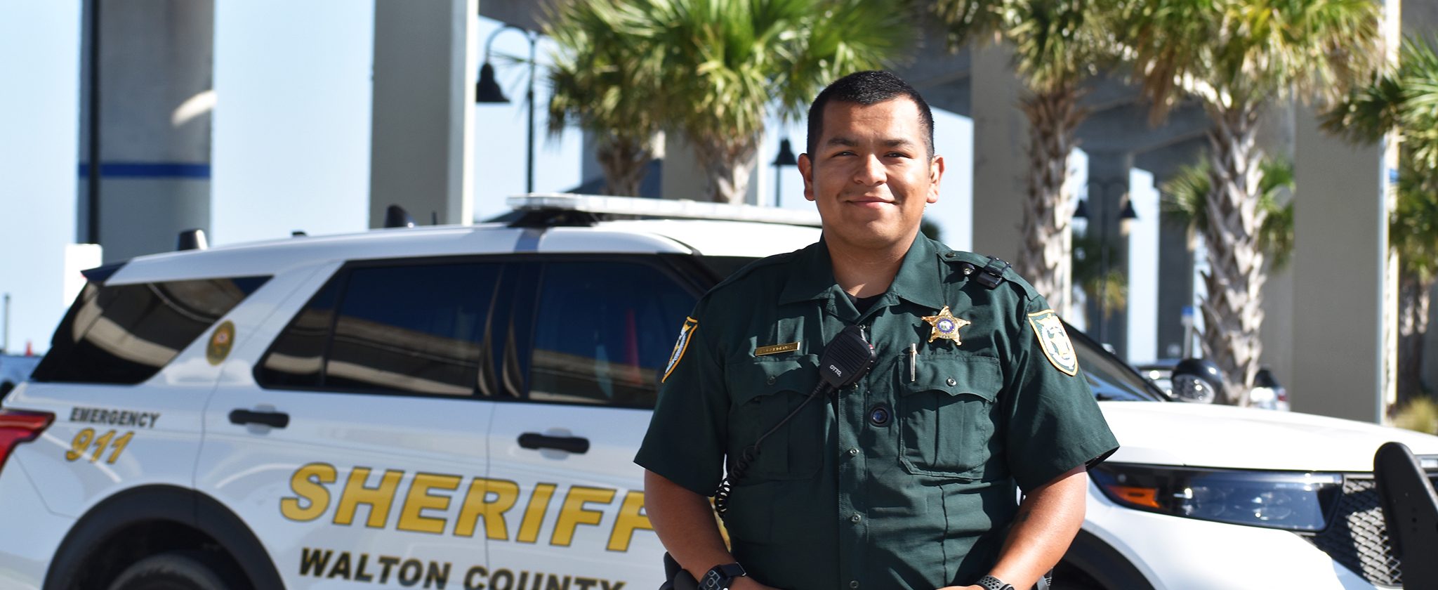 Hispanic male deputy standing in front of patrol car smiling
