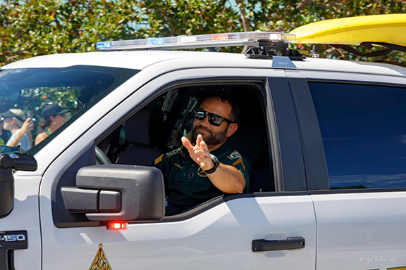 White male deputy with dark brown hair and dark brown beard waving out of patrol truck