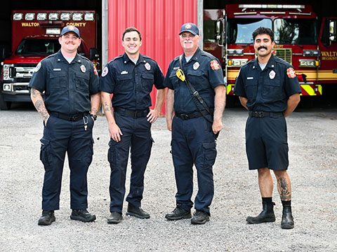Four male firefighters wearing navy blue uniforms standing in front of engine and ambulance