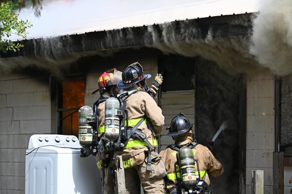 Firefighters in the doorway of a home on fire with heavy smoke billowing out