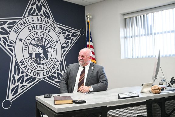 White male with a gray bear wearing a gray suit, white shirt, and burgundy tie sitting at desk smiling