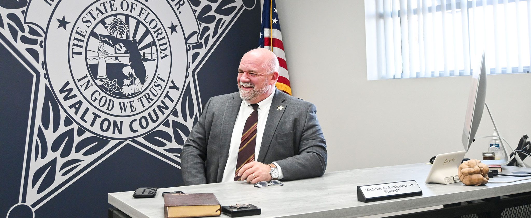 White male with bald head and gray beard sitting behind desk smiling