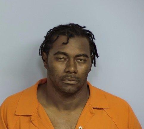 A man wearing an orange prison jumpsuit is facing the camera with a neutral or slightly tired expression. He has short dreadlocks and a medium-dark complexion, and the background is a plain light gray.