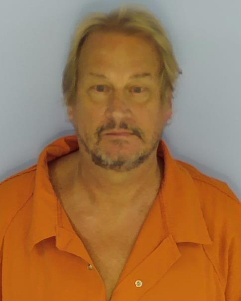 A white male with blond hair and a blank stare wearing an orange jumpsuit standing in front of a light blue wall.