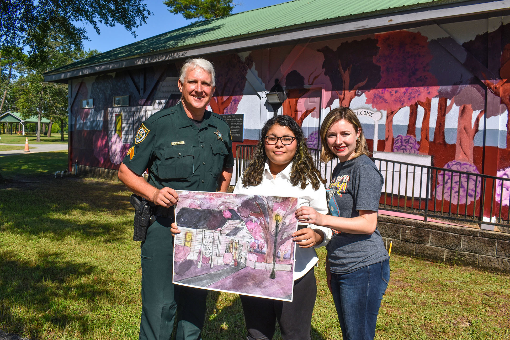White male deputy with white hair wearing a green sheriff's uniform standing with a female teacher and female art student in front of mural the art student painted