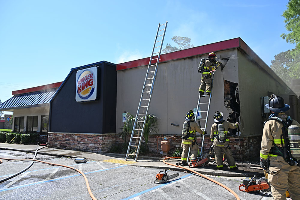 Firefighters on ladder making a large hole in the exterior wall of a Burger King restaurant