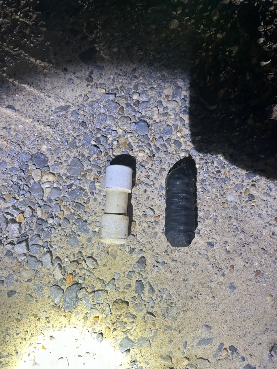Two objects are lying on a gravel and sand surface, illuminated by a flashlight. On the left is a segmented white cylindrical object made of stacked parts, and on the right is a black, ribbed cylindrical object with a rounded tip.