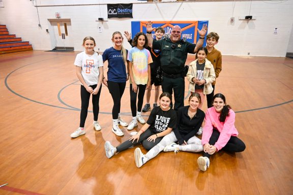 White male school resource deputy wearing a green sheriff's uniform, standing with highschoolers smiling